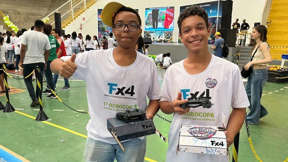 Two students holding Fx4 robots during an Instituto Amigos Droids robotics tournament, demonstrating robotics, programming, teamwork, and entrepreneurship in a hands-on learning journey